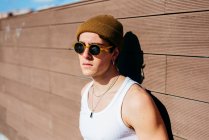 Hombre guapo joven moderno en gafas de sol de moda y gorro sombrero y camiseta blanca de pie cerca de la pared marrón en el día soleado en la calle de la ciudad - foto de stock