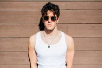 Modern young handsome man in trendy sunglasses and white tank top standing near brown wall on sunny day on city street — Stock Photo