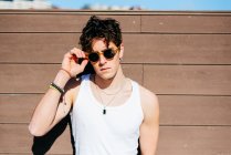 Modern young handsome man in trendy sunglasses and white tank top standing near brown wall on sunny day on city street — Stock Photo