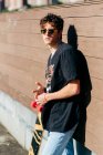 Bello hipster maschio in occhiali da sole in piedi vicino allo skateboard nella giornata di sole sulla strada della città — Foto stock