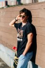 Bonito hipster masculino em óculos de sol em pé perto de skate no dia ensolarado na rua da cidade — Fotografia de Stock