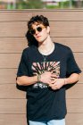 Modern young handsome man in trendy sunglasses and black t-shirt standing near brown wall on sunny day on city street — Stock Photo