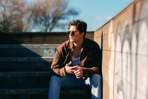 Giovane bel ragazzo in abiti casual guardando lontano mentre seduto sulle scale nella giornata di sole sulla strada della città — Foto stock