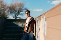Jeune beau mec en vêtements décontractés regardant la caméra tout en se tenant sur les escaliers par une journée ensoleillée sur la rue de la ville — Photo de stock