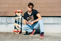 Beau mâle hipster dans des lunettes de soleil assis sur des hanches près de planche à roulettes sur une journée ensoleillée sur la rue de la ville — Photo de stock