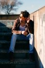 Young handsome guy in casual clothes touching ear and looking away while sitting on stairs on sunny day on city street — Stock Photo