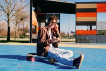 Joyeux hipster en vêtements décontractés souriant et naviguant smartphone tout en étant assis sur le skateboard et en écoutant de la musique sur le terrain de sport — Photo de stock