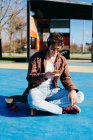 Happy hipster em roupas casuais sorrindo e navegando smartphone enquanto sentado no skate e ouvindo música em campo de esportes — Fotografia de Stock