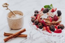 Crêpes savoureuses aux fraises et mûres fraîches décorées de feuilles de menthe placées sur une assiette près de bâtonnets de cannelle et d'une tasse de crème glacée sur une table en marbre — Photo de stock