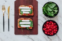 Top view of bread toasts with green spinach and mozzarella cheese on tomato sauce composed on wooden board with bowls of ingredients and cutlery on marble surface — Stock Photo