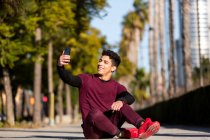 Веселий молодий етнічний хлопець у спортивному одязі посміхається і приймає селфі, сидячи на шляху під час фітнес-тренувань у сонячний день у місті — стокове фото