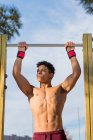 Low angle of muscular ethnic sportsman looking away and doing pull ups while hanging on bar against cloudy sky — Stock Photo