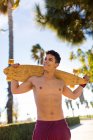Feliz chico hispano llevando longboard en hombros y mirando hacia otro lado mientras está de pie sobre un fondo borroso de terraplén en un día soleado en la ciudad - foto de stock