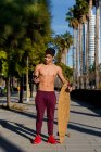 Полнотелый мускулистый этнический парень с длинной доской, улыбающийся и просматривающий смартфон, стоя на городской улице в солнечный день — стоковое фото
