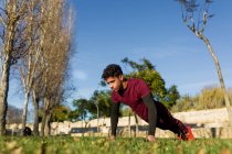 Junger hispanischer Mann in Sportbekleidung macht Liegestütze auf grünem Gras beim Fitnesstraining an einem sonnigen Tag im Park — Stockfoto