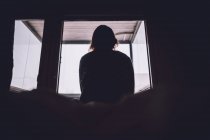 Из нижнего силуэта анонимной одинокой женщины, сидящей на кровати у окна в темной комнате дома — стоковое фото