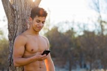 Glücklicher hispanischer Sportler ohne Hemd sitzt in der Nähe eines Baumstammes und surft während einer Pause im Fitnesstraining im Park am Smartphone — Stockfoto