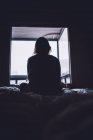 De silhueta abaixo de mulher solitária anônima sentada na cama contra a janela no quarto escuro em casa — Fotografia de Stock
