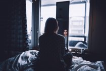 Вид сзади депрессивной молодой женщины, сидящей на кровати в темной спальне и смотрящей в зеркало дома — стоковое фото