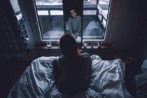Back view of depressed young female sitting on bed in dark bedroom looking at mirror at home — Stock Photo