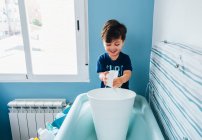 Sorrindo alegre menino brincando com água em balde branco se divertindo e segurando tigela na mão enquanto estava acima banheira de bebê no banheiro acolhedor — Fotografia de Stock