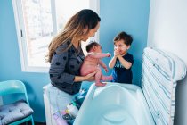 Adult caring woman in gently washing baby in baby bath in cozy bathroom while little son helping mom and holding bowl of warm water in hands — Stock Photo
