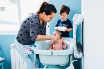 Adult caring woman in gently washing baby in baby bath in cozy bathroom while little son helping mom and holding bowl of warm water in hands — Stock Photo