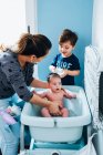 Femme attentionnée adulte en lavant doucement bébé dans le bain de bébé dans la salle de bain confortable tandis que le petit fils aide maman et tenant bol d'eau chaude dans les mains — Photo de stock
