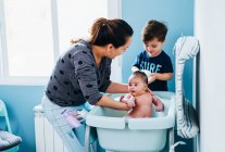 Adulto mulher carinhosa em suavemente lavar o bebê no banho de bebê no banheiro acolhedor, enquanto o pequeno filho ajudando a mãe e segurando tigela de água morna nas mãos — Fotografia de Stock