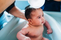 Femme attentionnée adulte en lavant doucement bébé dans le bain de bébé dans la salle de bain confortable tandis que le petit fils aide maman et tenant bol d'eau chaude dans les mains — Photo de stock