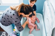 Erwachsene fürsorgliche Frau in sanft waschen Baby in Babywanne im gemütlichen Badezimmer, während kleiner Sohn hilft Mama und hält Schüssel mit warmem Wasser in den Händen — Stockfoto