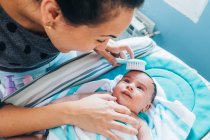 Erntefrau kämmt süßes Neugeborenes sanft in blaue Decke gehüllt mit kleiner weißer Haarbürste nach dem Bad, während Baby Mutter sorgfältig anschaut und auf Wickeltisch liegt — Stockfoto
