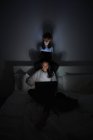 Focado menino e mulher adulta em sleepwear sentado cruzado pernas na cama e usando gadgets modernos, enquanto relaxa no quarto escuro em casa juntos — Fotografia de Stock