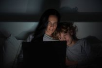 Glücklicher Junge, der sich auf die Schulter einer erwachsenen Frau lehnt, während er auf dem Bett liegt und nachts zu Hause zusammen Filme auf dem Laptop anschaut — Stockfoto