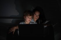Хлопчик спирається на плече дорослої жінки, лежачи на ліжку і дивитися фільм на ноутбуці вночі вдома разом — стокове фото