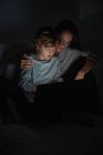 Мальчик, облокотившись на плечо взрослой женщины, лежа на кровати и смотря кино на ноутбуке ночью вместе дома — стоковое фото