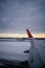 Wing of modern airplane on snowy terrain against dark forest at horizon and aircraft taking off from runway to gray sky in overcast weather at dusk in Norway — Stock Photo