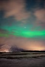 Pintoresco paisaje con asentamiento iluminado en la orilla del estrecho a pie de montañas nevadas bajo el cielo estrellado nublado con increíbles auroras boreales verdes en Lofoten - foto de stock