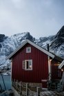 Casa de campo con paredes de rayas rojas y marcos de ventanas blancas en el muelle de madera en la orilla estrecha contra el municipio nevado en las estribaciones en invierno día nublado en Lofoten - foto de stock
