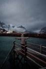 Back view of tourist in warm clothes and hood standing with arms outstretched on wooden pier against calm water of in fjord with township and snowy foggy mountains in Norway — Stock Photo