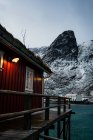 Cottage with red striped walls and white window frames on wooden pier on strait shore against snowy township at foothill in winter cloudy day in Lofoten — Stock Photo