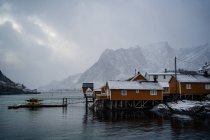 Casas de campo amarelas na costa do estreito contra cristas de montanha nevadas enevoadas em tempo nublado na Noruega — Fotografia de Stock