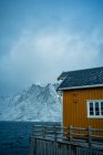 Casas de campo amarillas en la costa del estrecho contra las montañas nevadas brumosas en tiempo nublado en Noruega - foto de stock