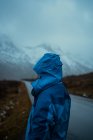 Vista trasera de una persona relajada irreconocible con ropa de abrigo azul y sudadera con capucha de pie en el camino de asfalto que va a las montañas nevadas de niebla en Lofoten - foto de stock
