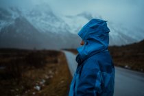 Повернення до розслабленої невпізнаваної людини в синьому теплому одязі та капюшоні, що стоїть на асфальтній дорозі, йде до засніжених туманних гір у Лофотені. — стокове фото