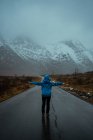 Вид на безликую женщину в голубой теплой одежде, наслаждающуюся идиллией и свежестью, стоя с протянутыми руками на асфальтовой дороге, идущей в заснеженные туманные горные хребты Норвегии — стоковое фото