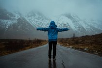 Vista posterior de la mujer sin rostro en ropa de abrigo azul disfrutando de idilio y frescura mientras está de pie con los brazos extendidos en el camino de asfalto que va a las cordilleras nevadas de niebla en Noruega - foto de stock