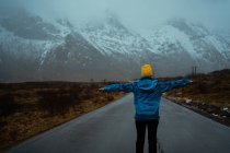Rückenansicht einer gesichtslosen Frau in blauer warmer Kleidung und leuchtend gelber Mütze, die Idylle und Frische genießt, während sie mit ausgestreckten Armen auf der Asphaltstraße steht, die in verschneite neblige Gebirgsketten in Norwegen führt. — Stockfoto