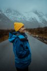 Vue latérale d'une femme heureuse et détendue vêtue de vêtements chauds bleus et d'un chapeau jaune vif profitant de la vie tout en regardant loin sur une route asphaltée allant vers des montagnes brumeuses enneigées à Lofoten — Photo de stock