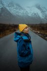 Vista lateral da mulher relaxada feliz em roupas quentes azuis e chapéu amarelo brilhante desfrutando da vida enquanto está de pé com os olhos fechados na estrada de asfalto indo para montanhas nebulosas nevadas em Lofoten — Fotografia de Stock